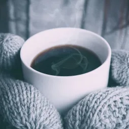 Close-up of a pair of hands wearing thick, gray knitted gloves holding a white ceramic cup filled with steaming coffee. The background is blurred, with a matching gray knitted sweater visible, creating a cozy and warm atmosphere. The steam rising from the coffee adds a sense of warmth and comfort, evoking feelings of relaxation and serenity on a cold day.