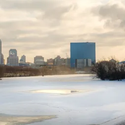 A frozen river with patches of open water reflecting sunlight stretches toward a city skyline in the background. The skyline features modern high-rise buildings, including a prominent blue glass tower. To the right, an industrial facility emits a large plume of white smoke. The sky is a mix of clouds and soft golden light, casting a wintery atmosphere over the scene.