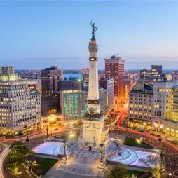 Aerial view of Monument Circle in Indianapolis, Indiana, at dusk. The Soldiers and Sailors Monument stands at the center, surrounded by historic and modern buildings with illuminated windows. The streets encircle the monument, showing streaks of light from moving traffic. The city skyline extends in the background, with a clear sky transitioning from blue to warm evening tones.